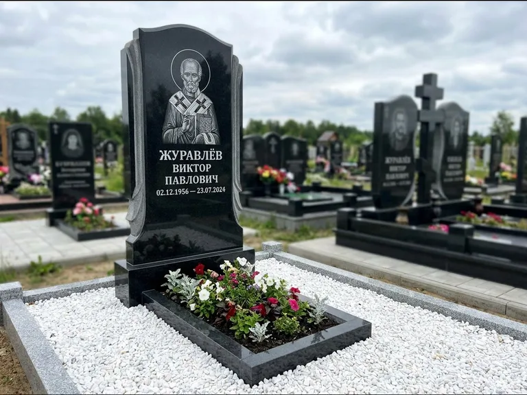 Памятник с гравировкой иконы: стела с образом в полный рост на обороте — гравировка образа Матроны Московской, низкие кусты спиреи в цвету, работа «Лебедева» во Владивостоке