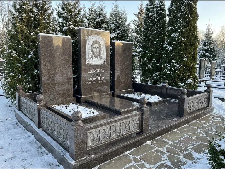 Памятник с гравировкой иконы: узкая вертикальная стела-пилон с ликом — икона в резной рамке-киоте, открытая лужайка без ограды, работа «Одинцов» во Владивостоке